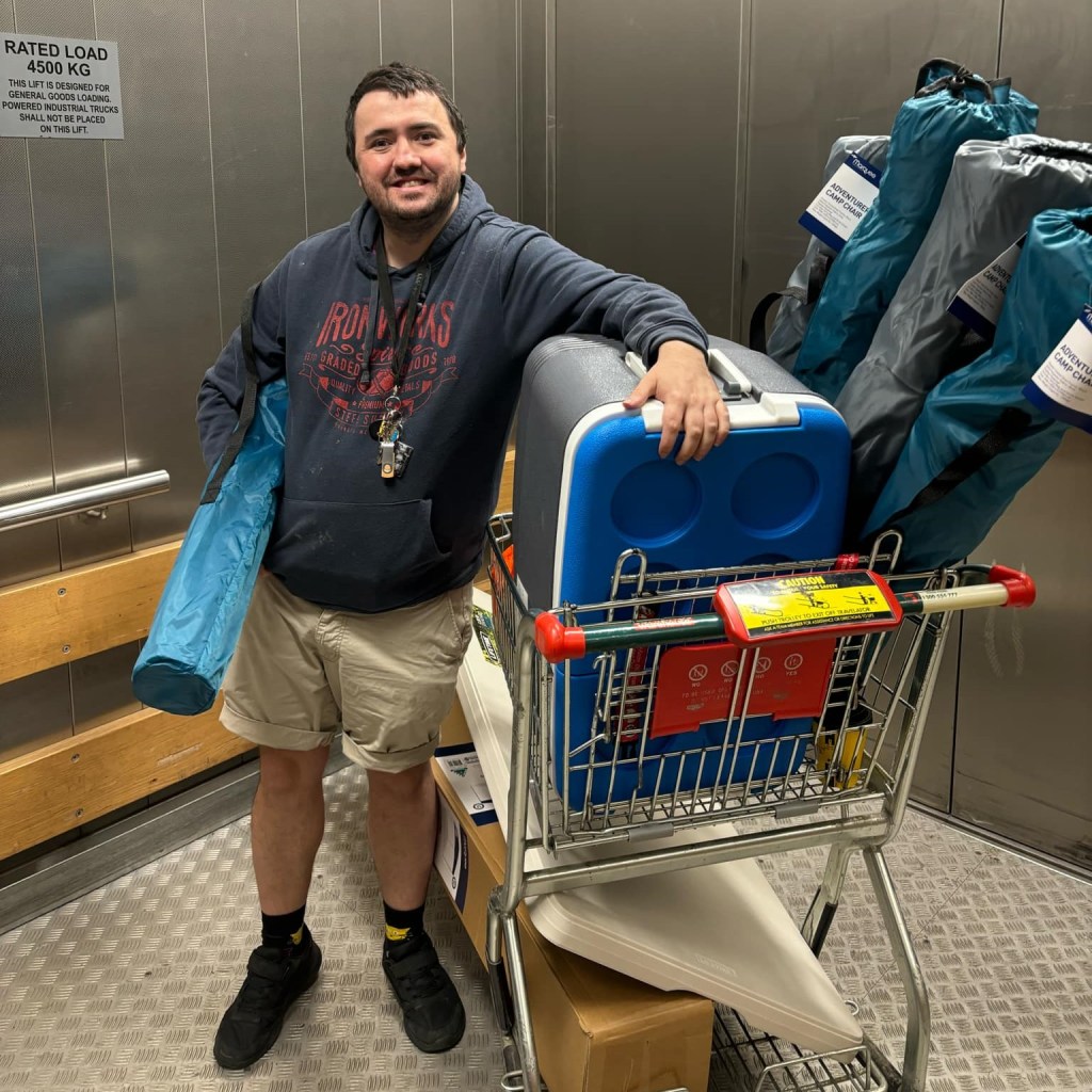 A smiling white man in his mid-late 30s with a cart full of camp chairs and a cooler. He looks to be purchasing them.