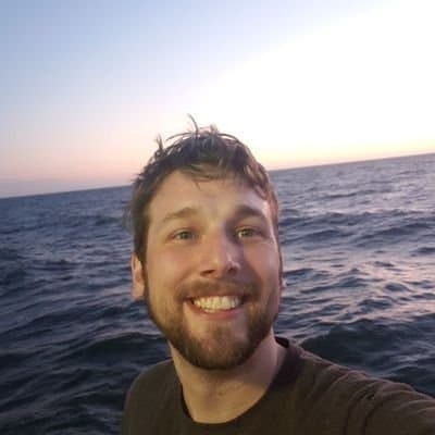 Shaun is a smiling white man in his 30s with grey eyes, brown hair, and a short beard against a backdrop of the ocean.
