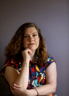 Tabitha is a lightly tanned woman with shoulder length brown curly hair, staring straight at the camera, with a serious look on her face. Her arms are half crossed with her chin resting on one curled hand. She has an ACAB tattoo on her wrist and she is wearing a multicolored dress. 